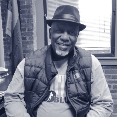 Black man in hat and vest sitting at a desk