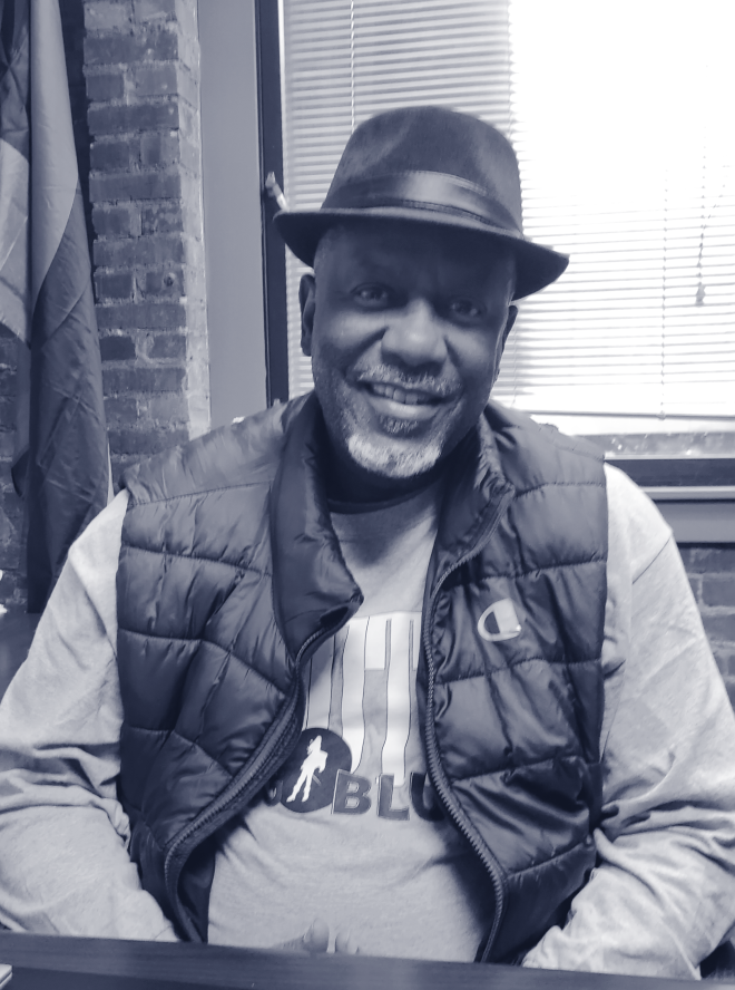 Black man in hat and vest sitting at a desk